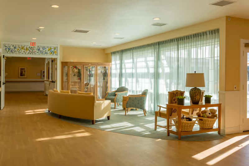 Bright senior living lounge with sofas and armchairs arranged by large windows with sheer curtains, and a side table holding a lamp and baskets.