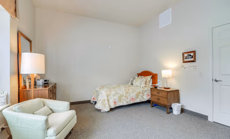 A cozy bedroom in an assisted living facility featuring a single bed with floral bedding, a wooden nightstand with a lamp and a small trash bin, a cushioned armchair with a pillow, a wooden dresser with a mirror, and a window with a beige curtain.