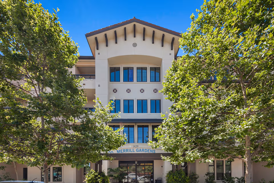 Exterior front view of Merrill Gardens at Willow Glen building with multiple floors, large windows, and a peaked roof. The entrance is framed by two large green trees on either side under a clear blue sky.
