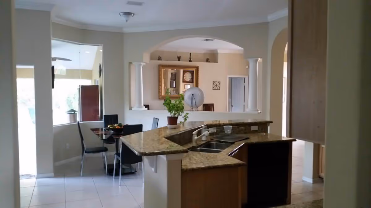 Interior view of a kitchen with a granite countertop island featuring a sink and faucet. In the background, there is a dining area with a glass table and black chairs, a potted plant on the counter, and decorative items including a large mirror and a clock on the wall.