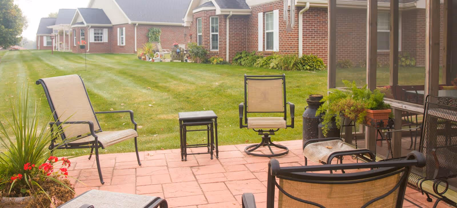Outdoor patio area with several metal-framed chairs with beige fabric seats and backs, a small black side table, potted plants, and a well-maintained grassy lawn extending towards a red brick building with white-trimmed windows in the background.