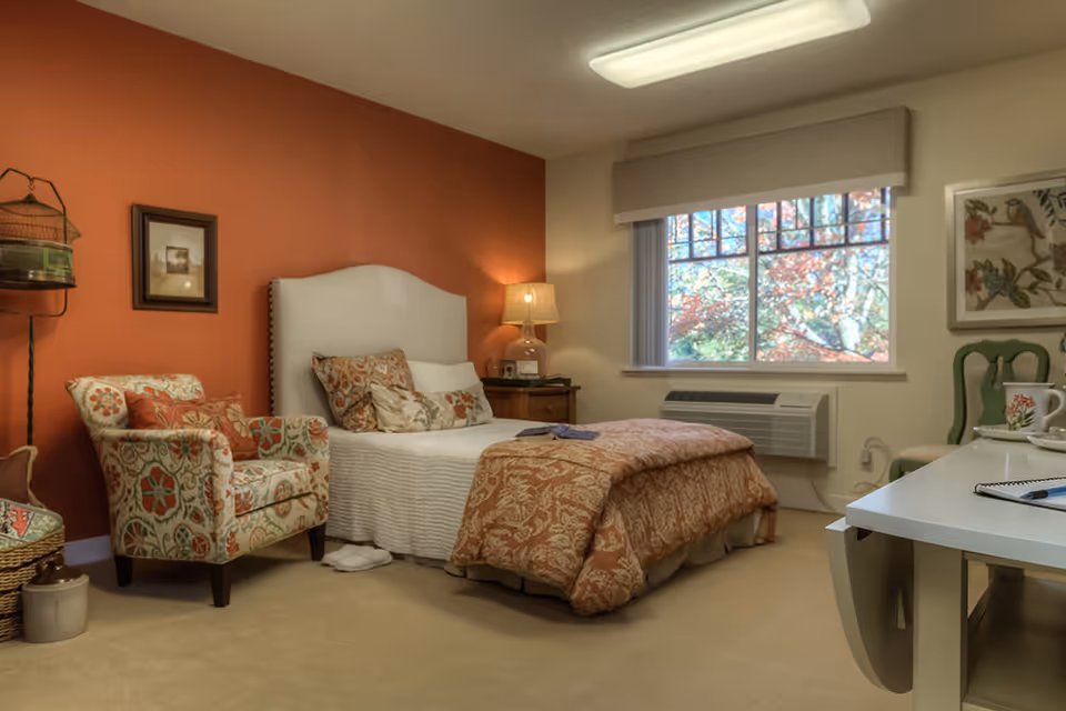 A cozy bedroom with a single bed featuring a white headboard and patterned bedding. There is a floral upholstered armchair next to the bed, a wooden nightstand with a lamp, and a window showing colorful autumn leaves outside. A white desk with a green chair and some items on top is visible on the right side of the room. The walls are painted in warm tones, with one accent wall in burnt orange.