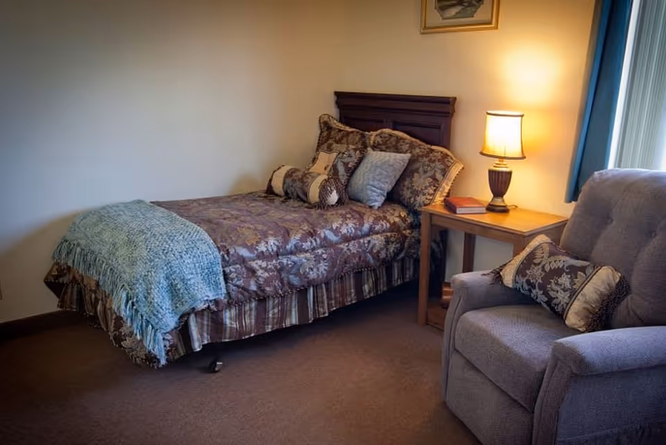 A cozy bedroom with a single bed covered in a floral patterned bedspread and multiple pillows. Next to the bed is a wooden nightstand with a lit table lamp and a book. A comfortable upholstered armchair with a decorative pillow is positioned nearby. A window with blue curtains allows natural light into the room.