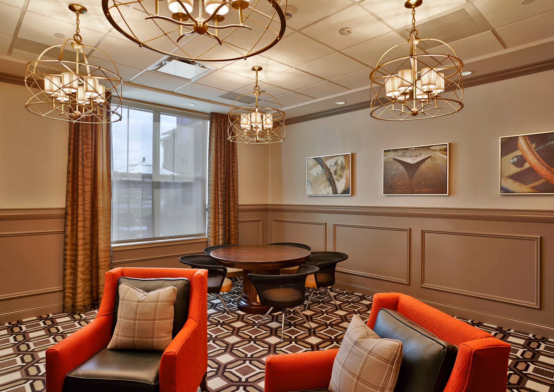 A cozy interior room with two orange armchairs featuring plaid and black cushions, a round wooden table with four black chairs, three modern gold chandeliers hanging from the ceiling, patterned carpet flooring, beige walls with decorative paneling, three framed abstract art pieces on the wall, and a large window with brown curtains.