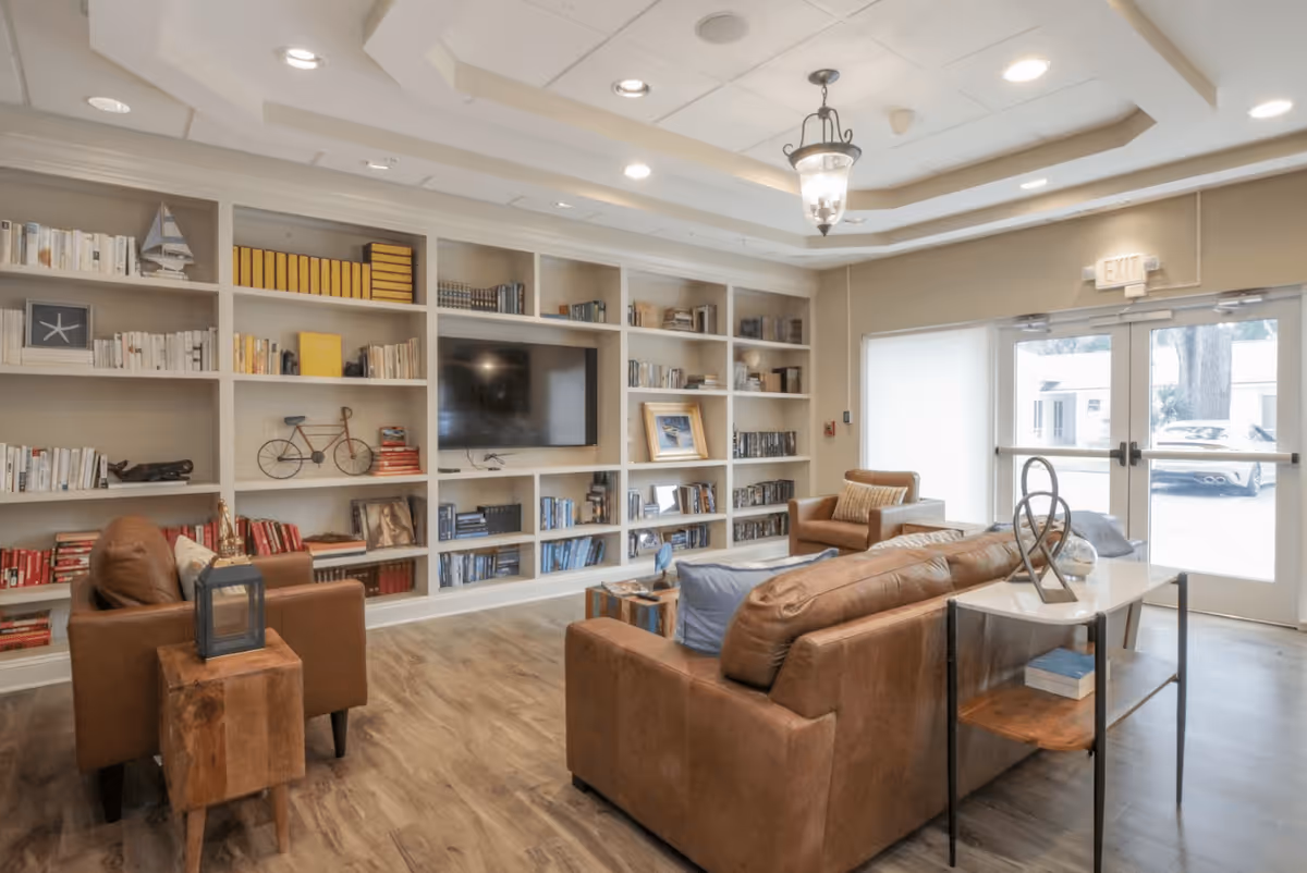 A cozy lobby area featuring brown leather sofas, a coffee table, and a large bookshelf filled with books. There is a television mounted on the wall and large windows allowing natural light to enter the space.