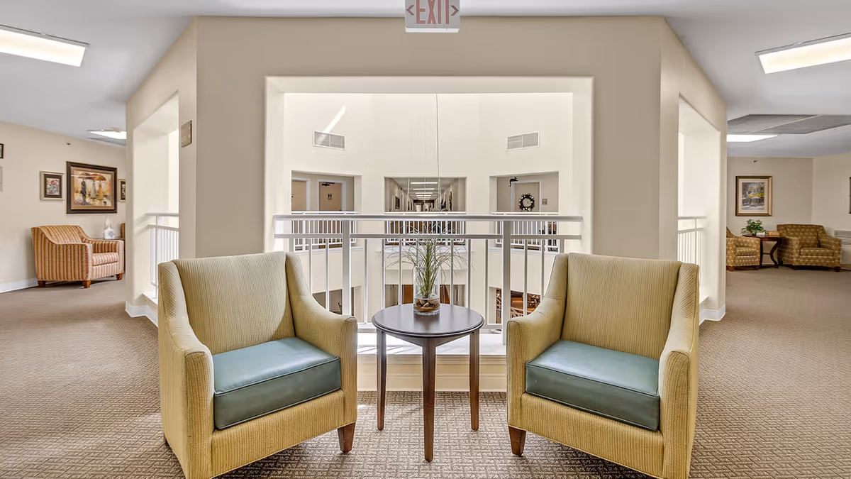 A seating area in a senior living facility with two beige armchairs featuring green cushions positioned on either side of a small round wooden table with a glass vase containing a plant. The area overlooks a hallway with railings and framed artwork on the walls. An exit sign is visible above the seating area.