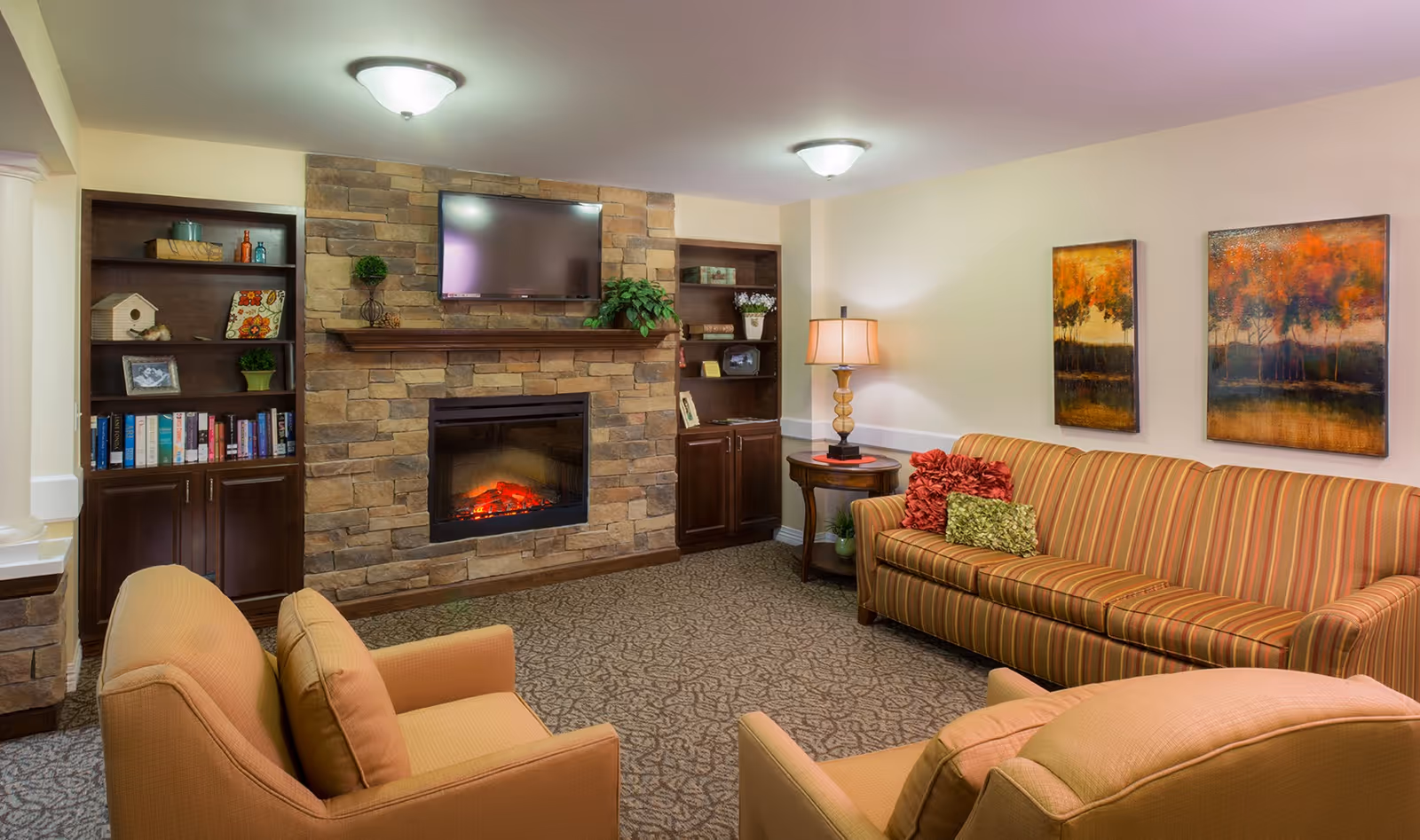 Cozy living room with a stone fireplace and a mounted flat-screen TV above it. The room features built-in dark wood shelves on either side of the fireplace, decorated with books, plants, and decorative items. There is a striped sofa with colorful pillows along one wall, two beige armchairs facing the fireplace, a side table with a lamp, and two autumn-themed paintings on the wall.