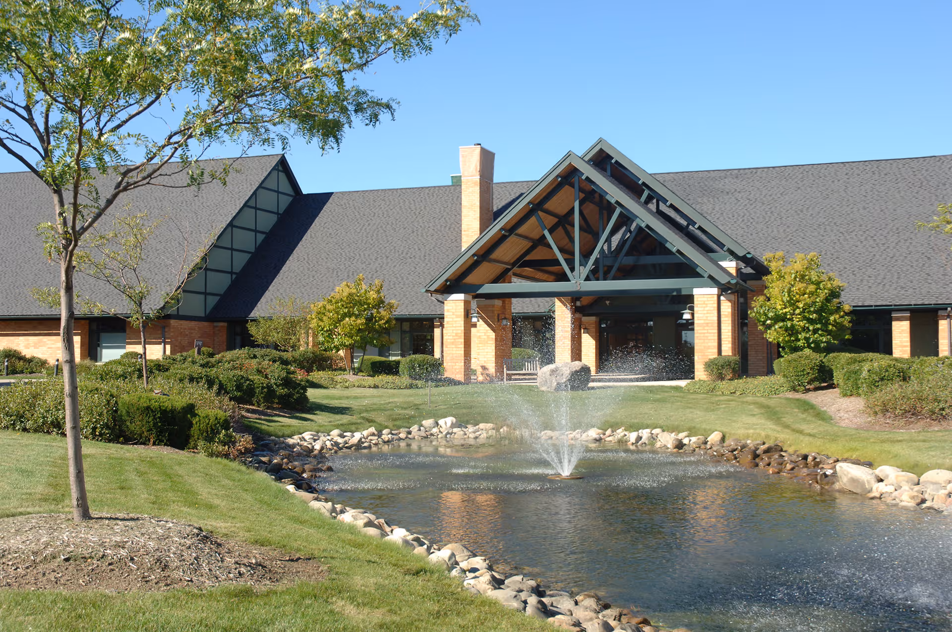 Exterior view of a single-story building with a large covered entrance supported by brick pillars. In front of the building is a landscaped area with green grass, bushes, small trees, and a pond with a water fountain in the center. The sky is clear and blue.