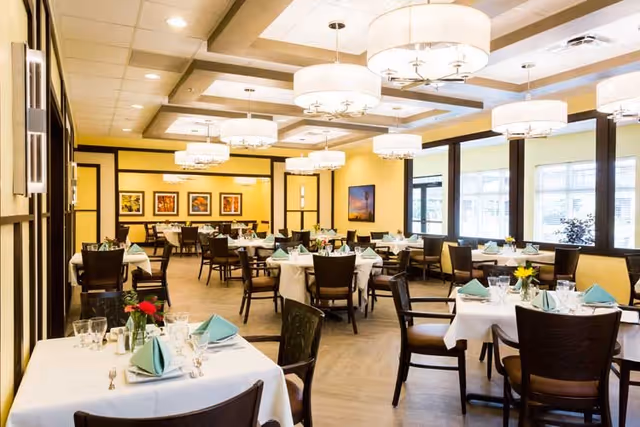 A bright and spacious dining room with multiple tables covered in white tablecloths, each set with folded green napkins, glassware, and silverware. The room features large windows allowing natural light, yellow walls with framed artwork, dark wooden chairs, and modern ceiling light fixtures.
