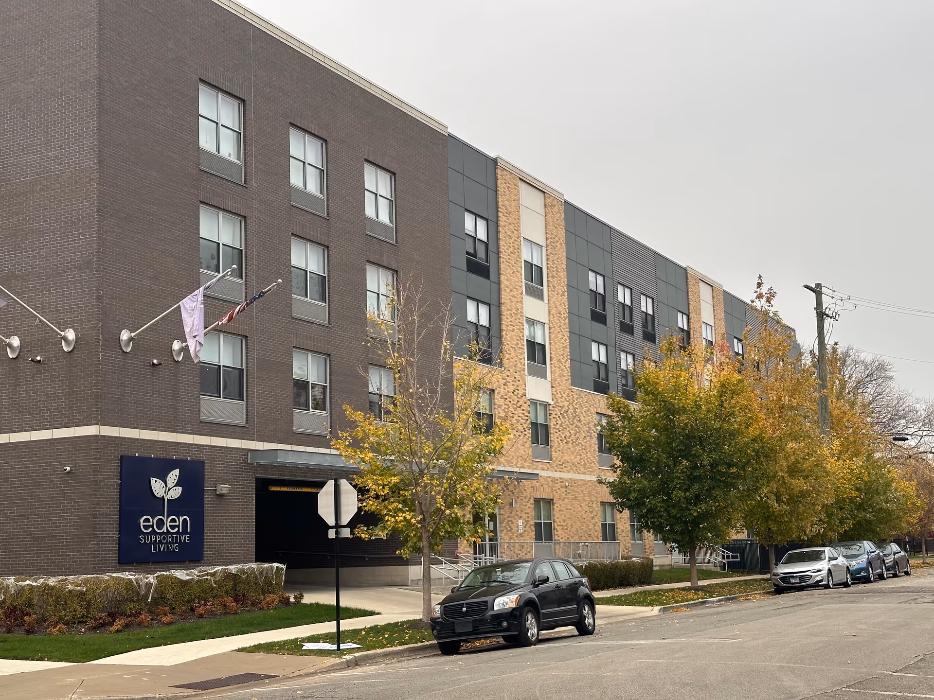 Exterior view of a multi-story building with a mix of dark brown and beige brick facade, several windows, and a sign that reads 'eden SUPPORTIVE LIVING'. There are a few trees with autumn foliage along the sidewalk and several parked cars on the street in front of the building under an overcast sky.