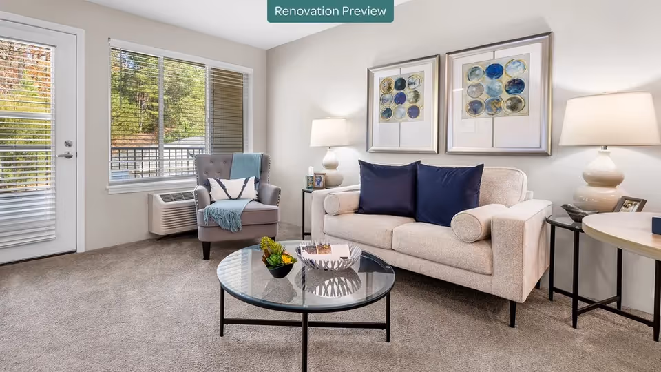 Bright furnished living room with a sofa, armchair, round glass coffee table, lamps, and wall art next to a window and glass door.
