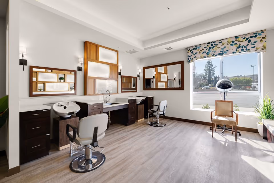 A bright and modern salon area with two styling chairs in front of large mirrors mounted on the wall. There are two hair washing sinks integrated into dark wood cabinetry. A large window with a floral valance lets in natural light, and a vintage hair dryer chair is positioned near the window. The room has light wood flooring and white walls with contemporary lighting fixtures.
