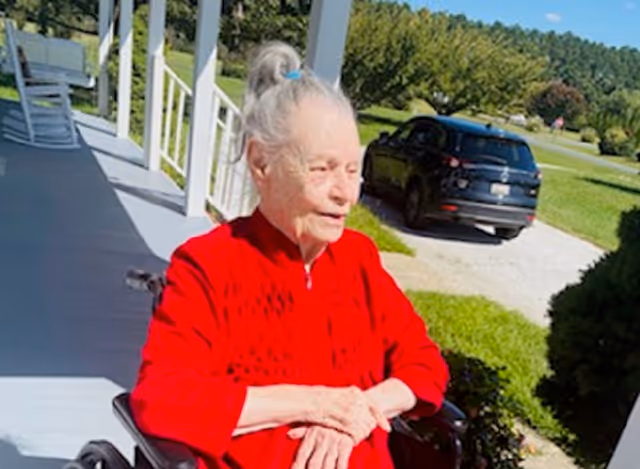 An elderly woman with white hair tied in a ponytail is sitting in a wheelchair on a porch. She is wearing a bright red sweater and looking to the side. Behind her, there is a black car parked on a driveway, green grass, trees, and a clear blue sky.