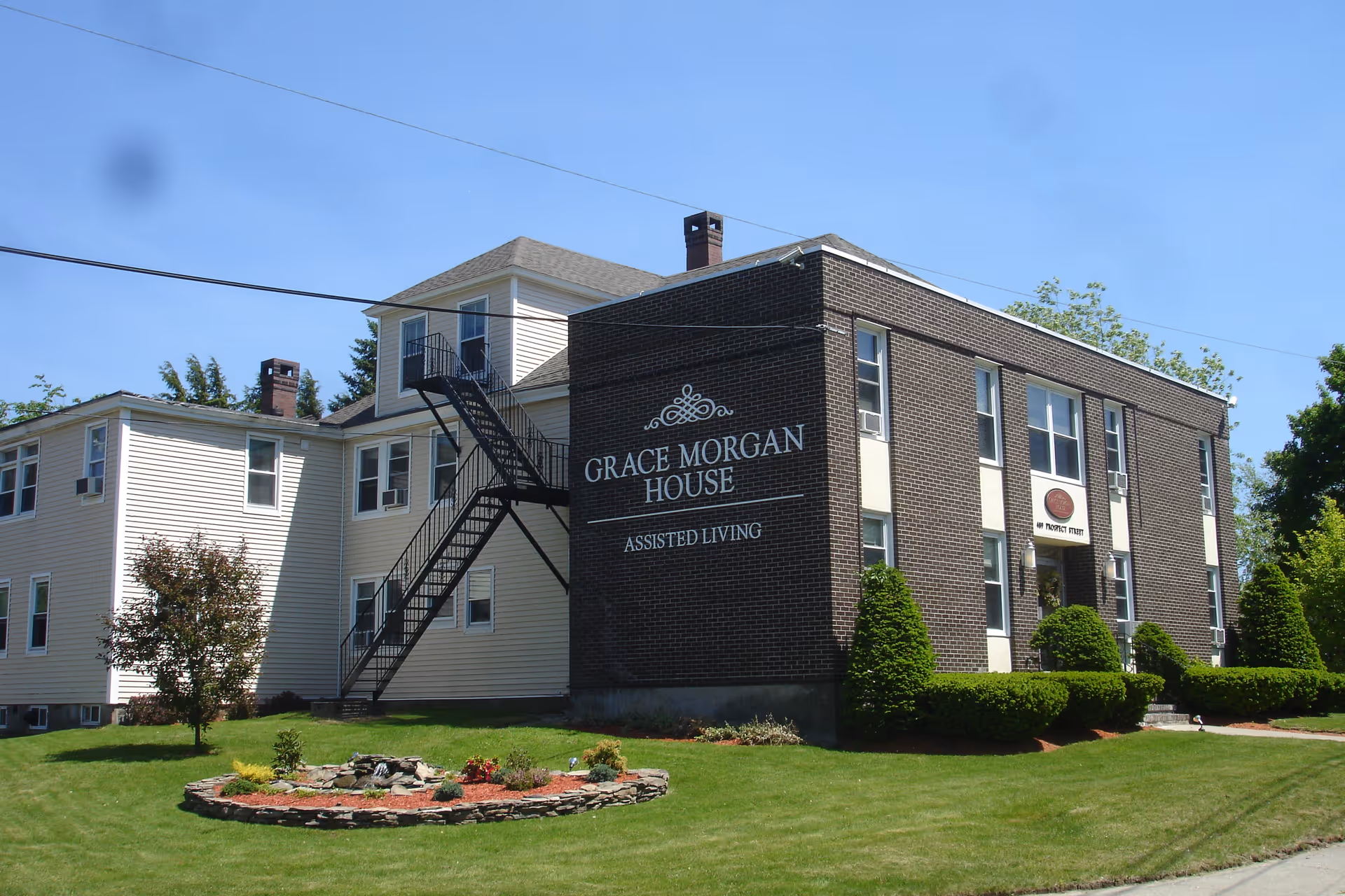 Exterior view of Grace Morgan House, an assisted living facility. The building has a combination of light-colored siding and dark brick walls, with multiple windows and an external black metal staircase. The front lawn is neatly maintained with green grass, bushes, and a small circular flower bed. The sky is clear and blue.