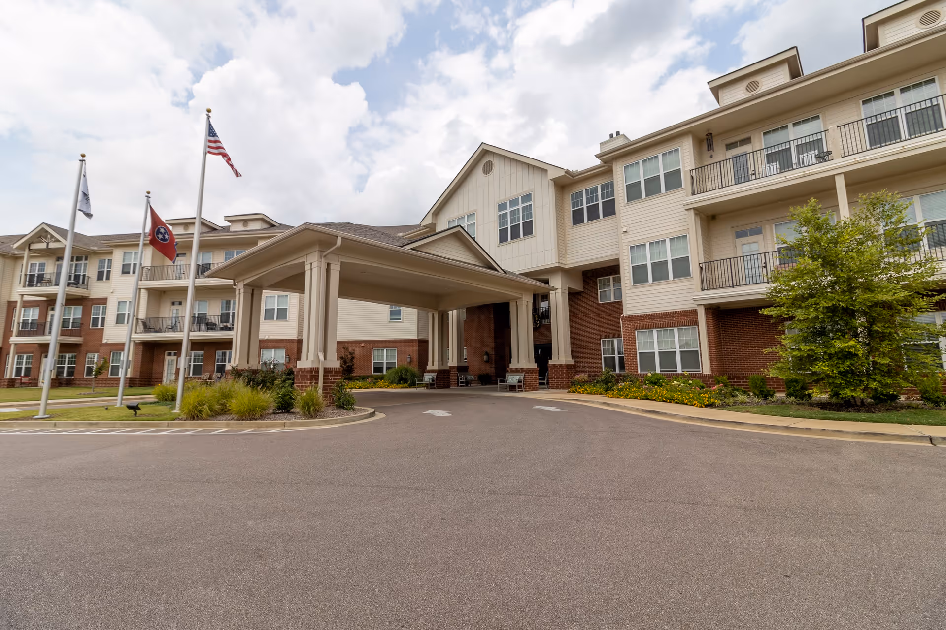 Exterior view of a multi-story senior living facility with a covered entrance driveway, three flagpoles with flags, and landscaped greenery around the building under a partly cloudy sky.