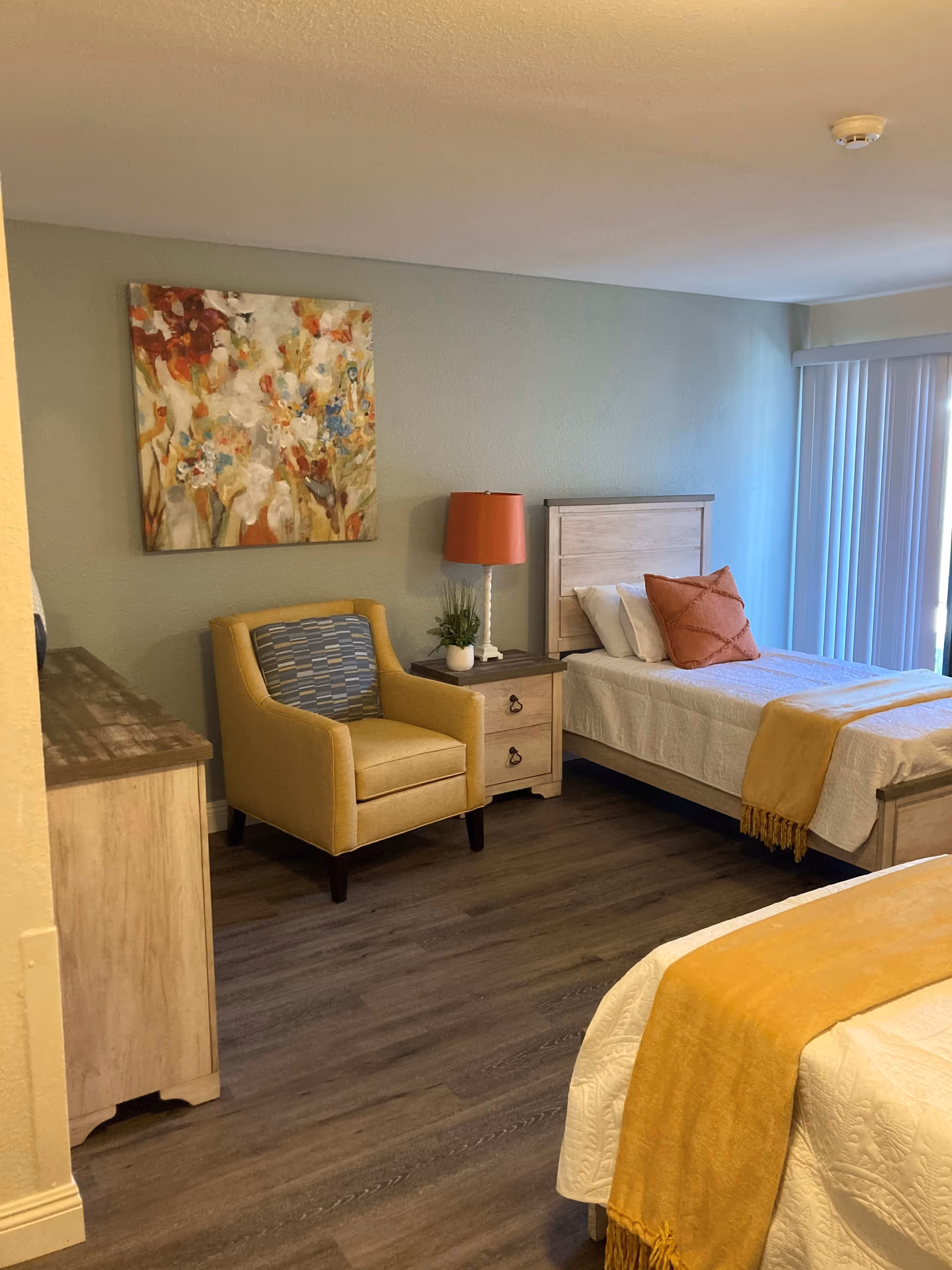 A bedroom with two single beds, each with white bedding and a yellow throw blanket. There is a wooden nightstand between the beds with a lamp and a small plant. A yellow armchair with a patterned cushion is next to the nightstand. A colorful abstract floral painting hangs on the wall above the armchair. The room has wood flooring and vertical blinds covering a window or sliding door.