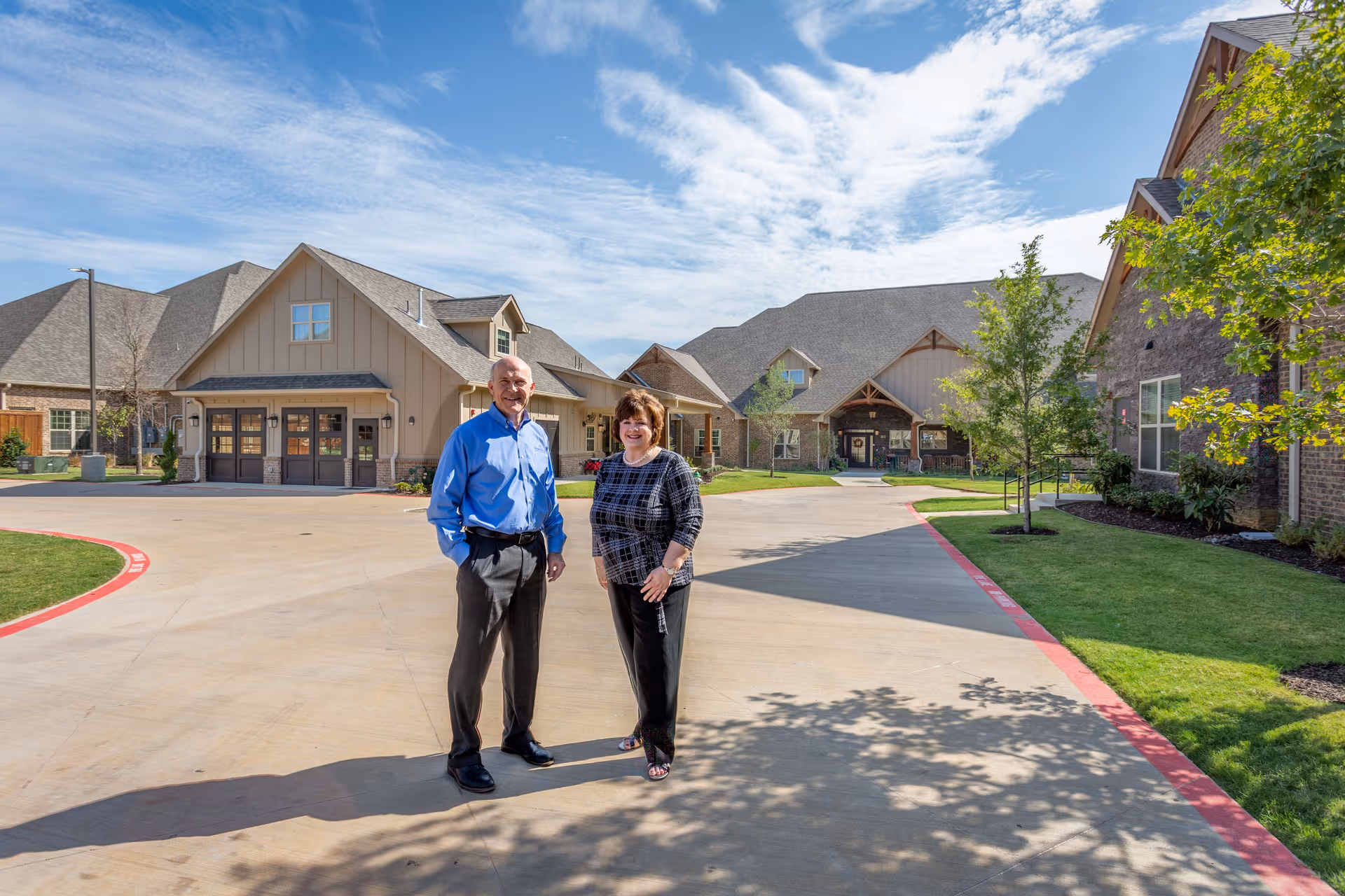 Two people standing outside on a paved driveway in front of a large assisted living facility building with multiple peaked roofs, surrounded by green lawns and trees under a partly cloudy blue sky.