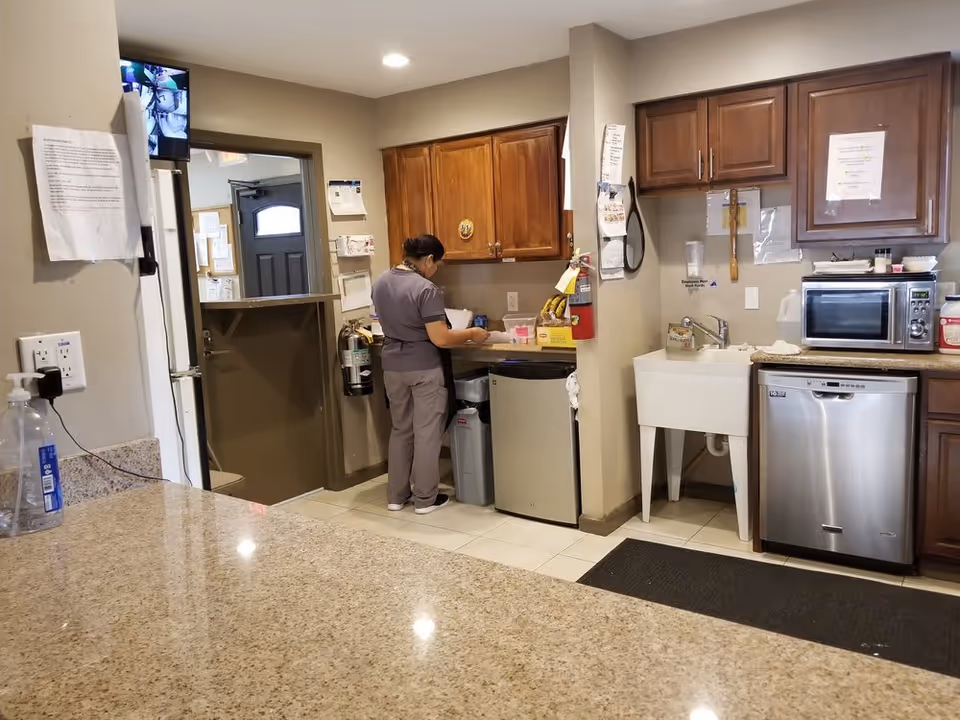 A person in a gray uniform is standing in a kitchen area preparing something on the counter. The kitchen has wooden cabinets, a small refrigerator, a dishwasher, a microwave, a utility sink, and various items on the counters. There is a granite countertop in the foreground and a door in the background with a security camera monitor mounted on the wall.
