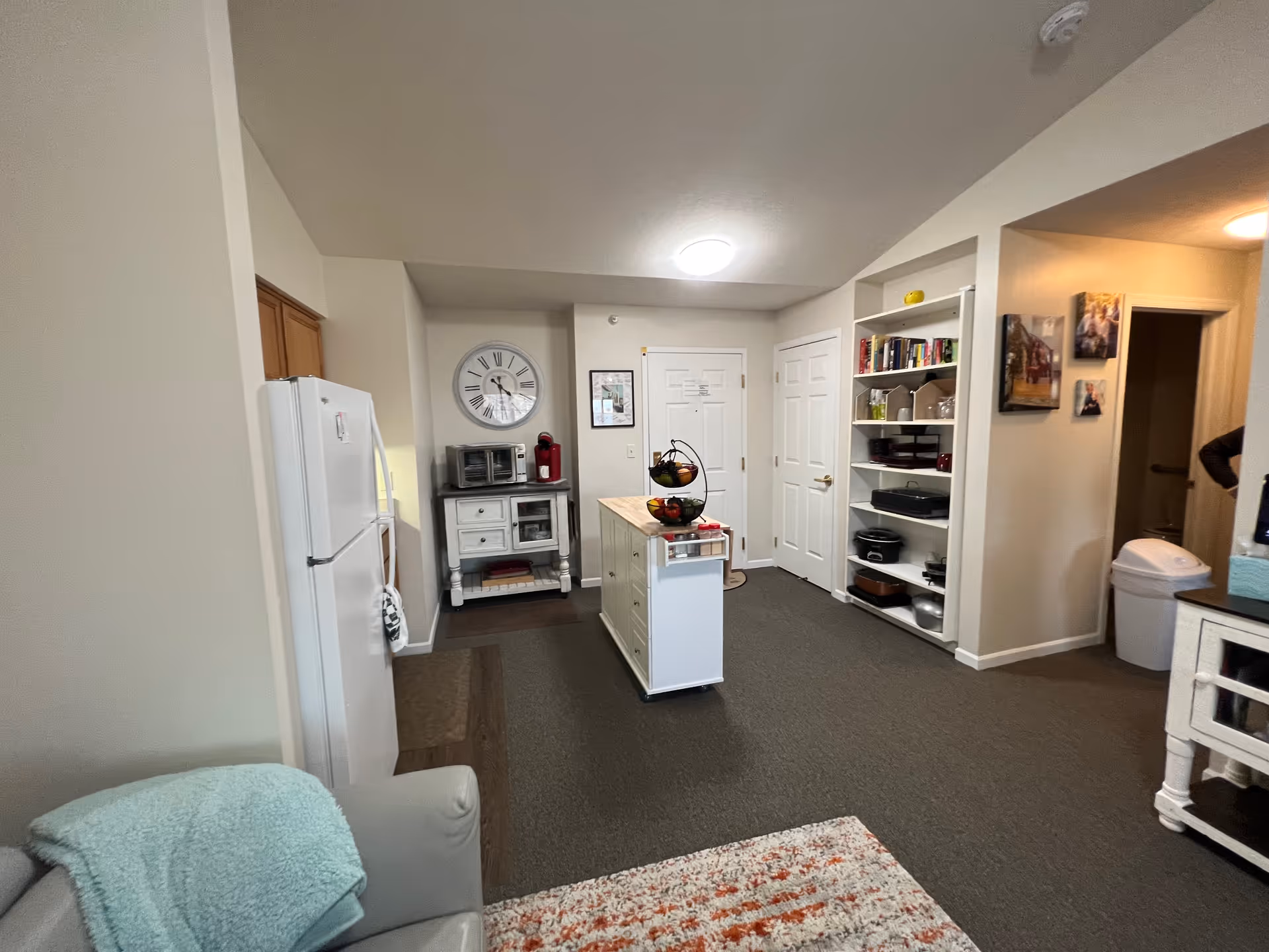 Interior view of a senior living facility kitchen and common area. The image shows a white refrigerator, a small kitchen island with a fruit basket, a wall clock, a microwave, and shelves with books and kitchenware. There is a gray armchair with a light blue blanket in the foreground and a carpeted floor with a patterned rug.