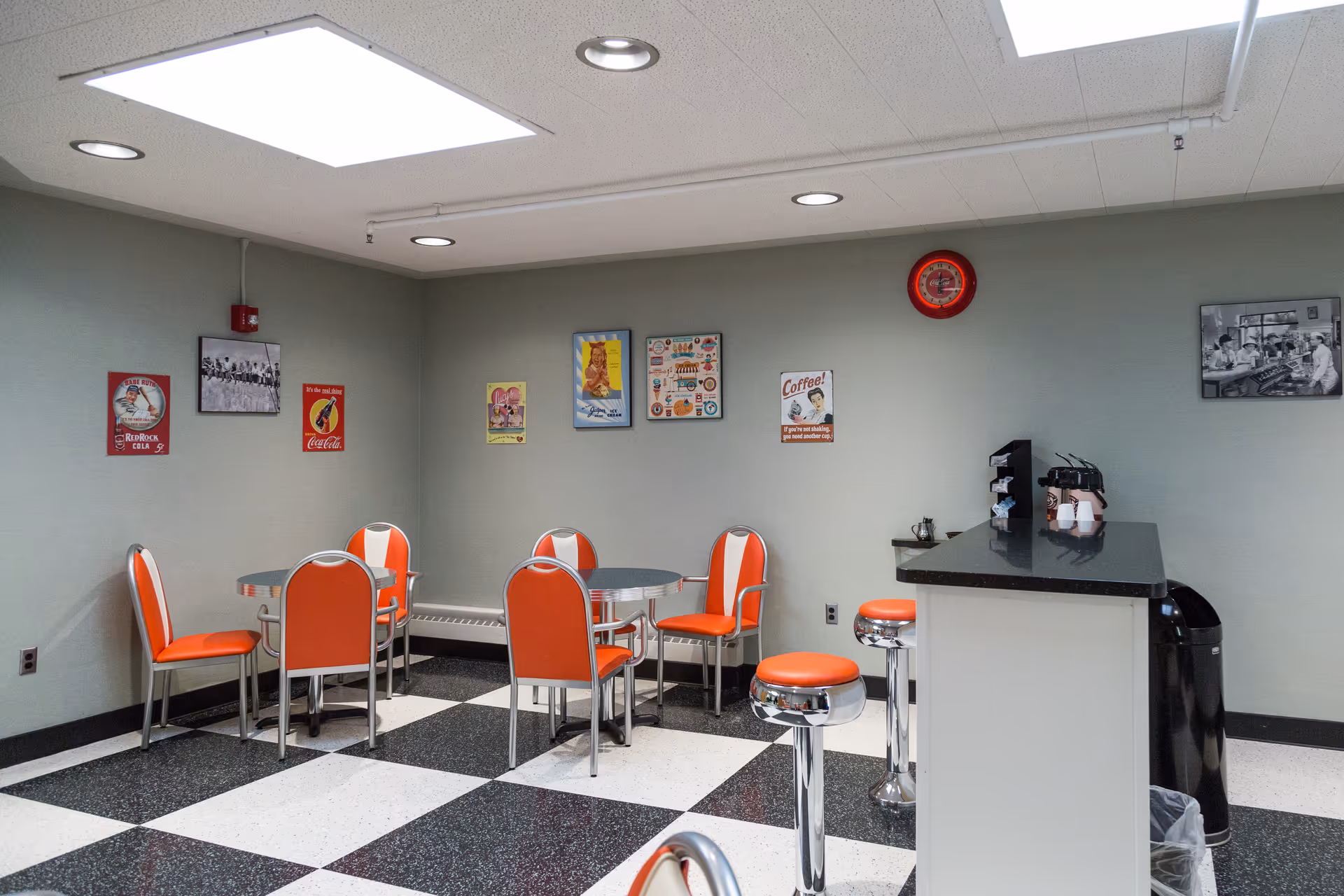 A retro-style dining area with orange and white chairs around small round tables and matching bar stools at a counter. The floor has a black and white checkered pattern, and the walls are decorated with vintage posters and a red clock. The ceiling has recessed lighting and a skylight.