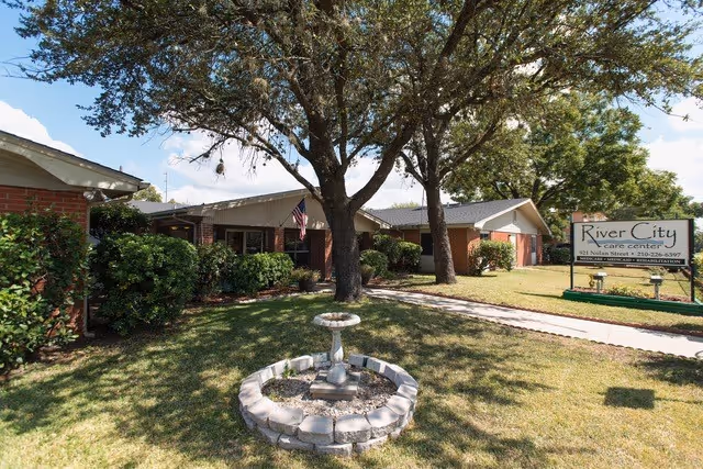 Exterior view of River City Care Center showing a single-story brick building with a well-maintained lawn, trees, and a small circular stone-bordered garden feature with a birdbath in the foreground. A sign with the facility's name and contact information is visible on the right side of the image.