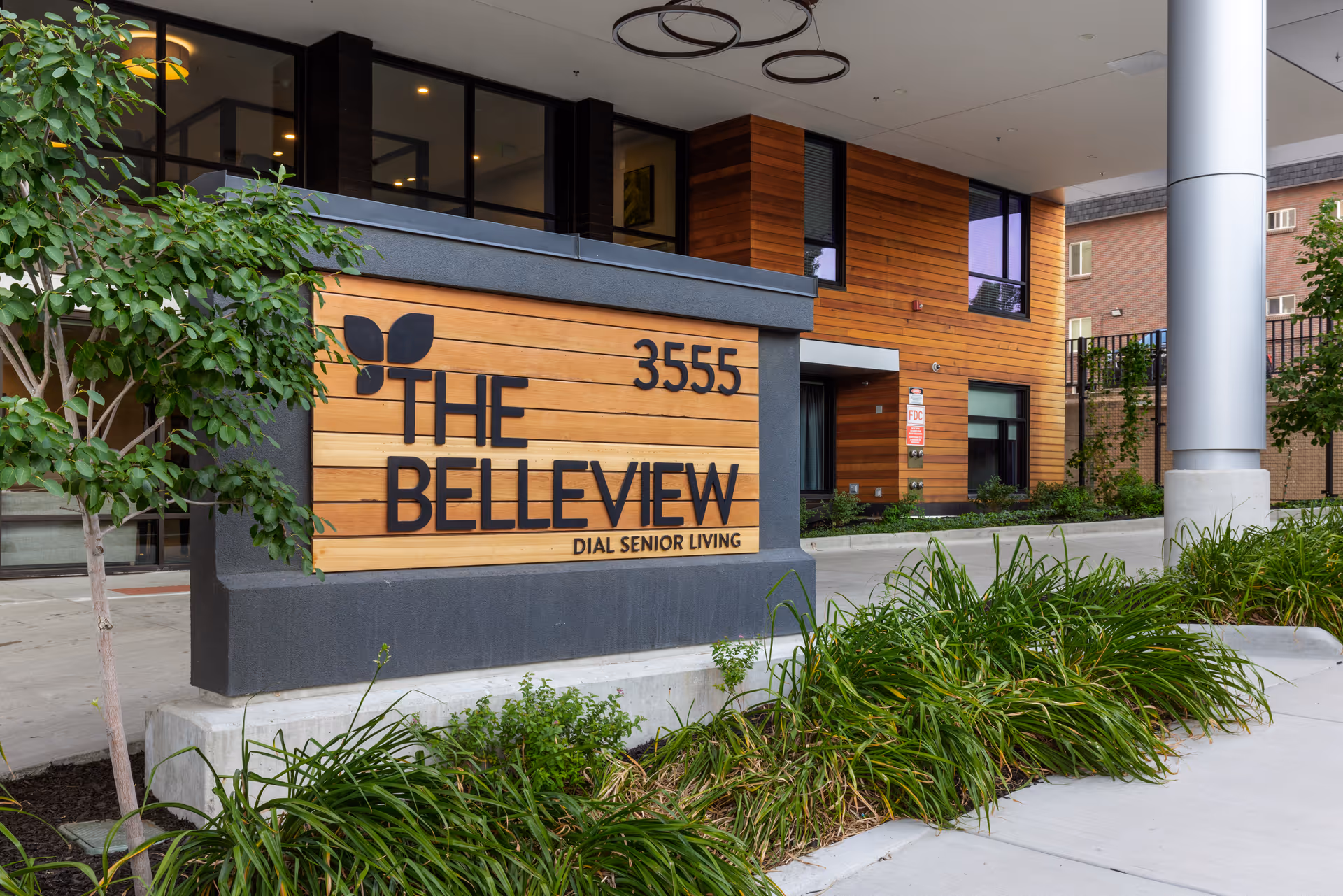 The entrance sign for The Belleview Dial Senior Living facility, featuring a wooden panel with black lettering and a leaf logo, surrounded by greenery and part of the building visible in the background.