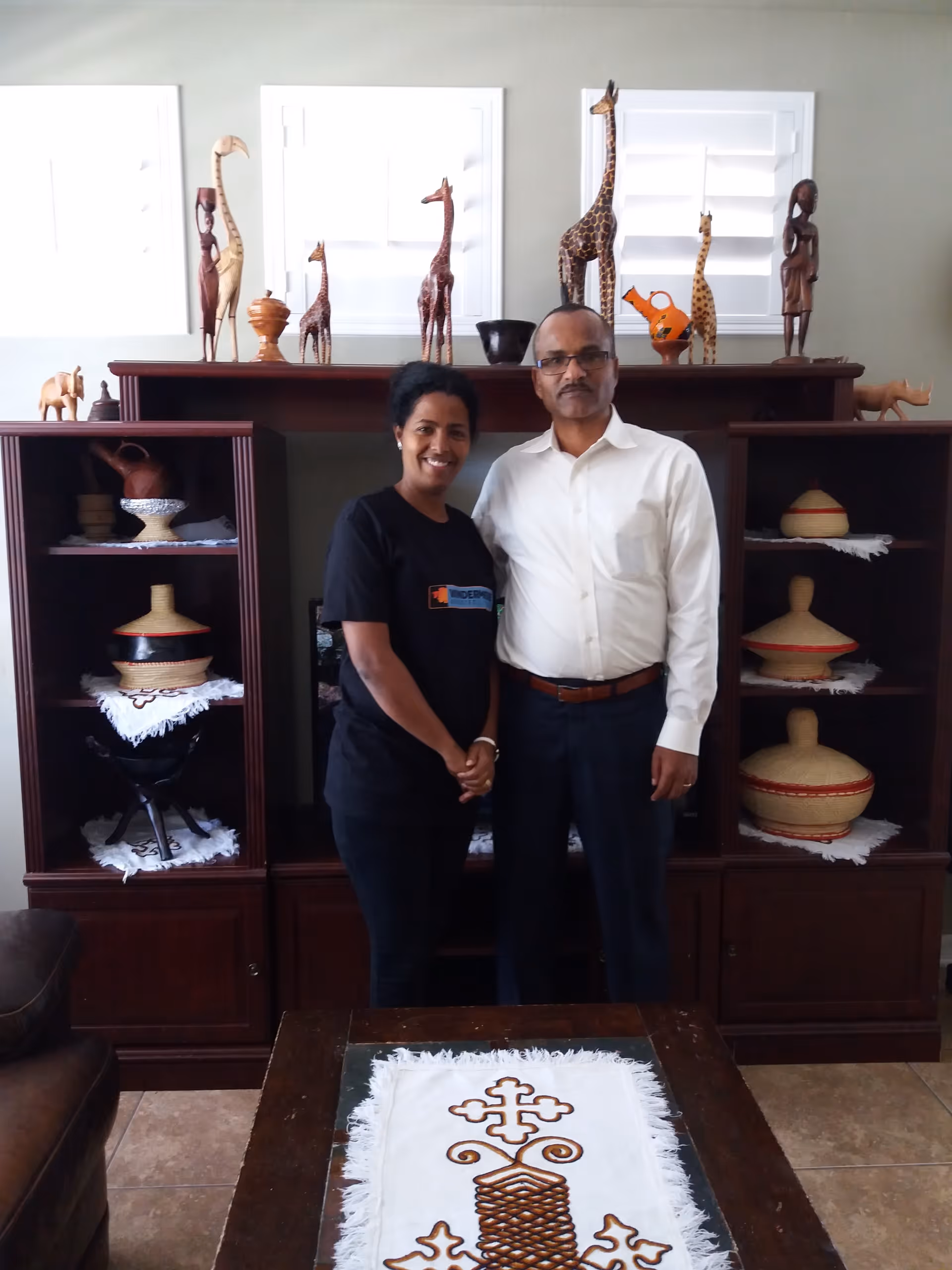 A man and a woman standing together in a living room area with wooden shelves behind them displaying various decorative items including giraffe figurines and traditional baskets. The woman is wearing a black t-shirt and the man is wearing a white shirt and dark pants.