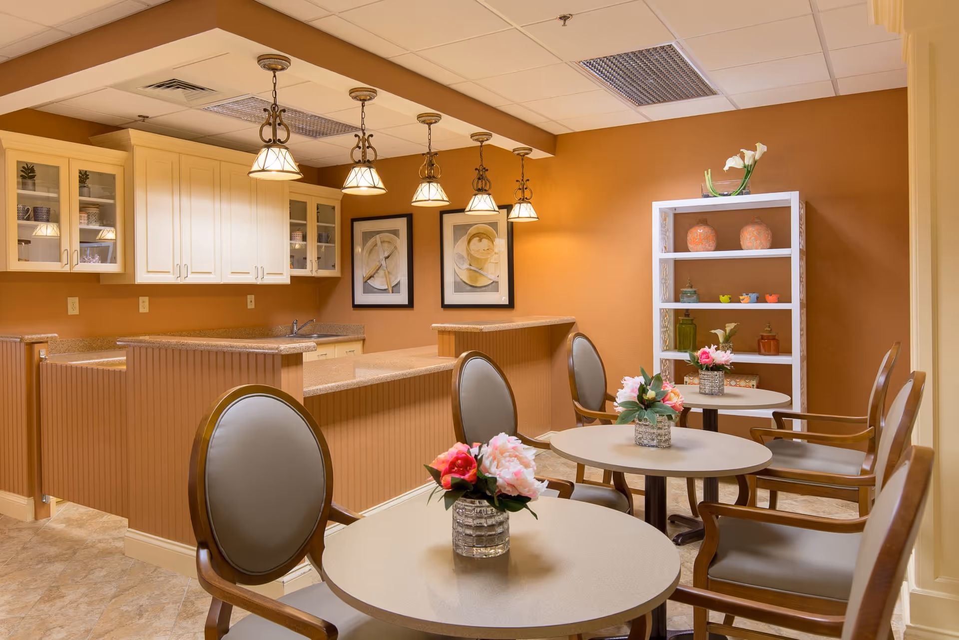 A cozy dining area in a senior living facility with round tables, each adorned with a small vase of pink and red flowers. The space features cushioned chairs with wooden frames, a kitchen counter with beige cabinets and granite countertops, and warm orange walls decorated with framed artwork and a white shelving unit holding decorative items.