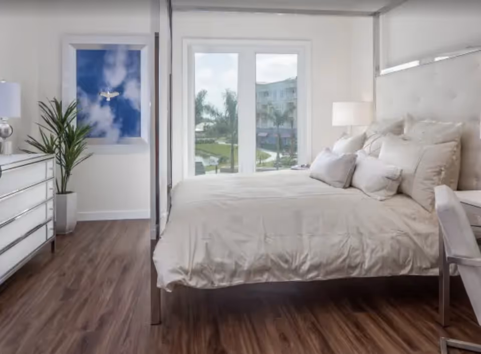 Bright and modern bedroom with a large bed covered in beige bedding and multiple pillows, a white dresser with a lamp and a potted plant on the left, a framed picture of a bird flying in a blue sky on the wall, and large windows showing a view of palm trees and a building outside.