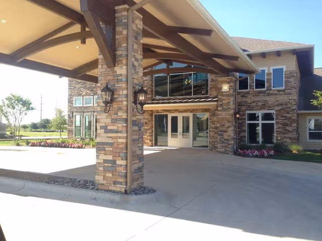 Entrance of a senior living facility with a covered driveway supported by stone pillars, large glass doors, and windows on a brick and stone building exterior. There are outdoor lantern-style lights mounted on the pillars and flower beds near the building.
