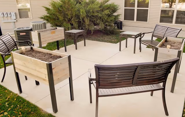 Outdoor patio area with metal benches and small tables arranged on a concrete surface, surrounded by raised wooden planters and greenery near a building with windows.