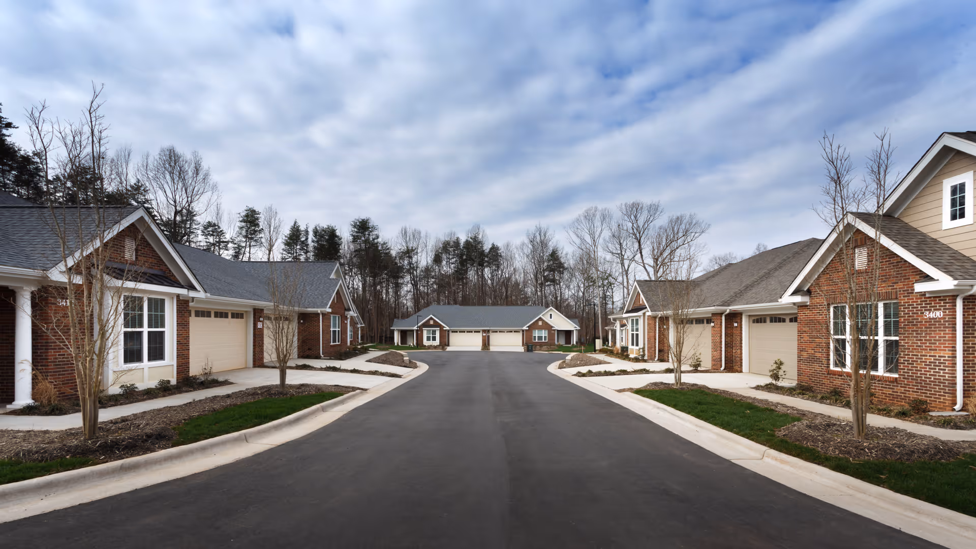 A paved road flanked by single-story brick residential buildings with garages, small trees, and landscaped areas under a partly cloudy sky.