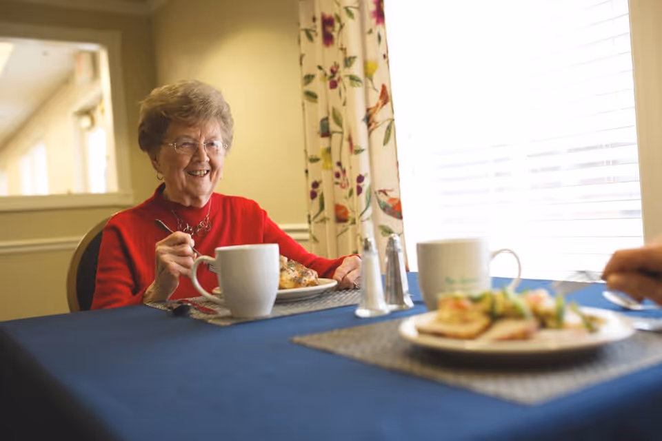 An older woman in a red sweater smiling while seated at a dining table with coffee mugs and plates of food.