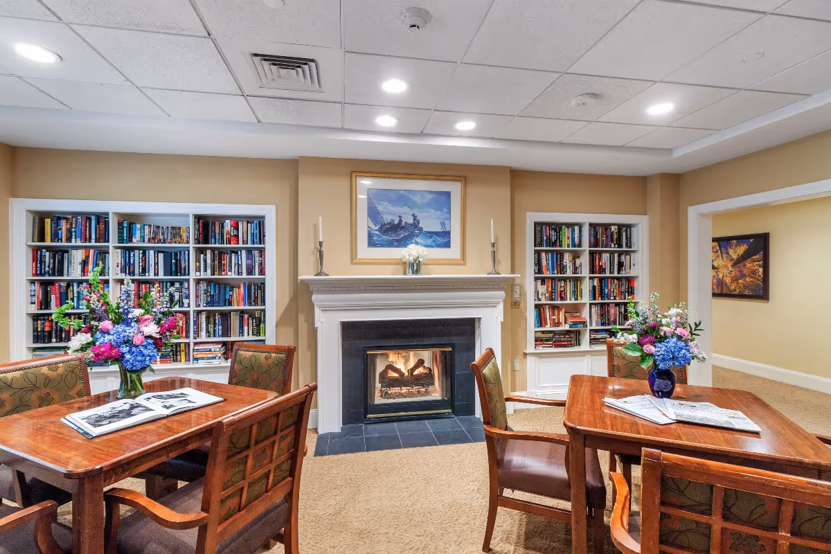 Cozy interior room with a lit fireplace centered between two built-in bookshelves filled with books. Two wooden tables with floral arrangements and chairs are placed in front of the fireplace. The walls are painted beige, and there is a framed painting above the fireplace. The room has recessed ceiling lights and carpeted flooring.