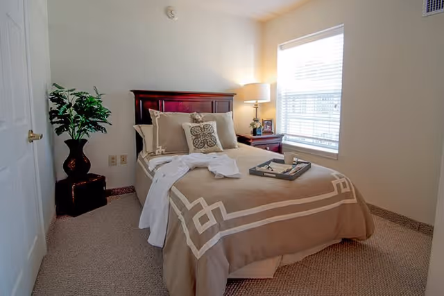 A cozy bedroom with a neatly made bed featuring beige and white bedding, a wooden headboard, and decorative pillows. A tray with a cup and book rests on the bed. There is a bedside table with a lamp and a framed photo next to a window with blinds. A potted plant is placed in the corner near a closed door.