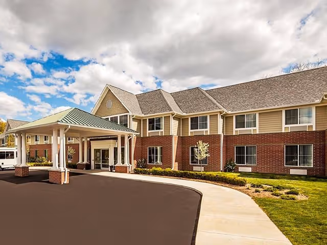 Exterior view of a two-story senior living facility building with a covered entrance, brick and beige siding, windows with blinds, a paved driveway, and a sidewalk surrounded by green grass and small trees under a partly cloudy sky.
