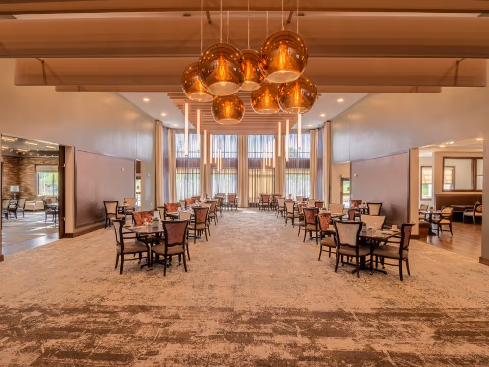 Spacious dining room in a senior living facility with multiple tables and chairs arranged neatly. Large windows with sheer curtains allow natural light to fill the room. Modern pendant lights hang from the ceiling, and the carpet has a patterned design.