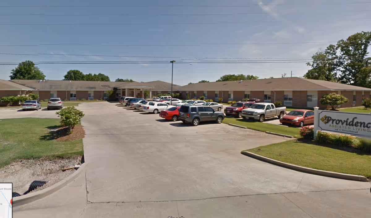 Exterior view of a single-story assisted living facility building with a parking lot in front filled with cars. There is a sign on the right side that reads 'Providence Assisted Living of Cleveland'. The building is surrounded by green grass and trees under a clear blue sky.
