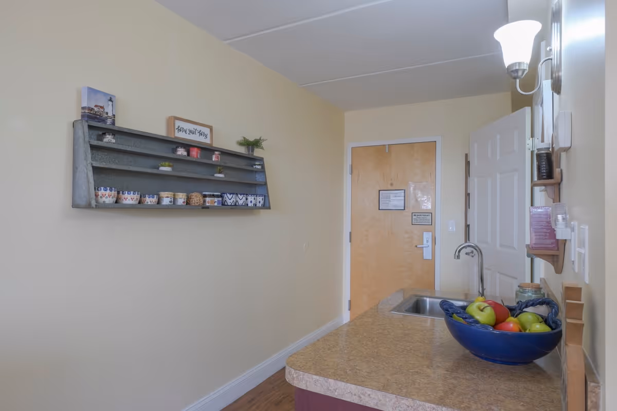 A small kitchen area with a countertop holding a blue bowl filled with apples. There is a sink with a faucet on the countertop. On the wall above the countertop, there are shelves holding cups, jars, and small decorative items. A wooden door is visible in the background.