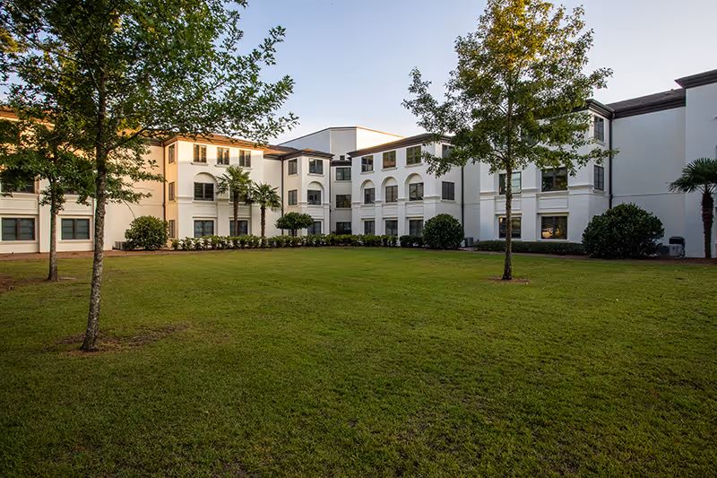 Well-maintained grassy courtyard with trees in front of a white multi-story senior living building.