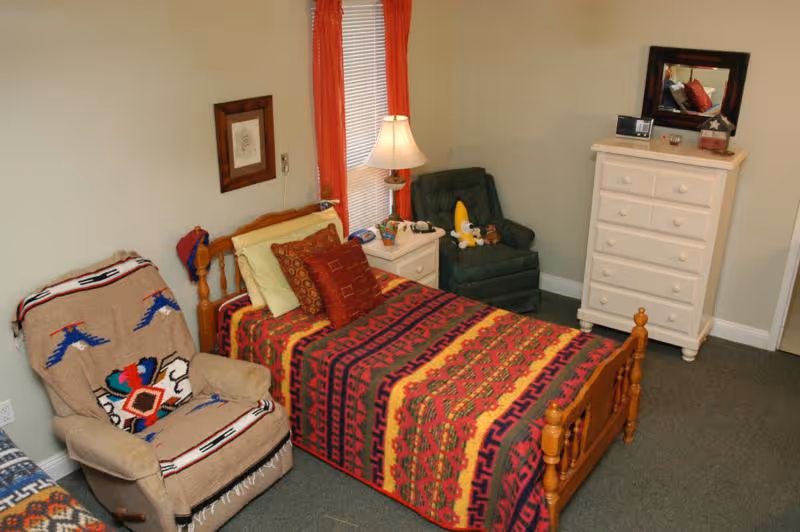 A cozy bedroom with a single wooden bed covered in a colorful patterned blanket and multiple pillows. Next to the bed is a white nightstand with a lamp, a phone, and some small items. There is a green armchair with stuffed toys on it and a beige recliner chair with a patterned throw blanket. A white dresser with drawers and a mirror on top is placed against the wall. The room has beige walls and a window with red curtains.