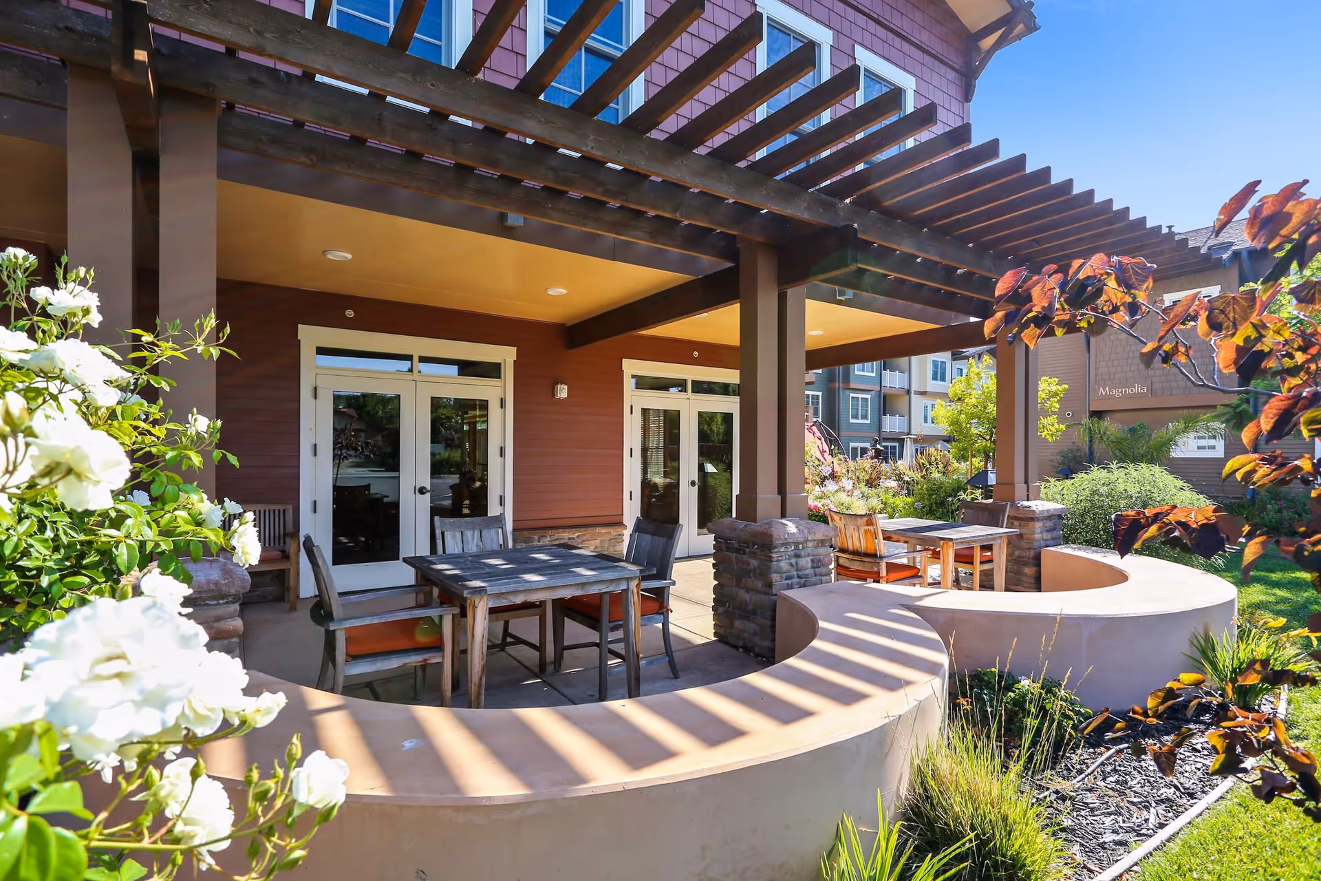 Covered outdoor patio with tables and chairs under a wooden pergola in front of the senior living building, surrounded by landscaped plants.