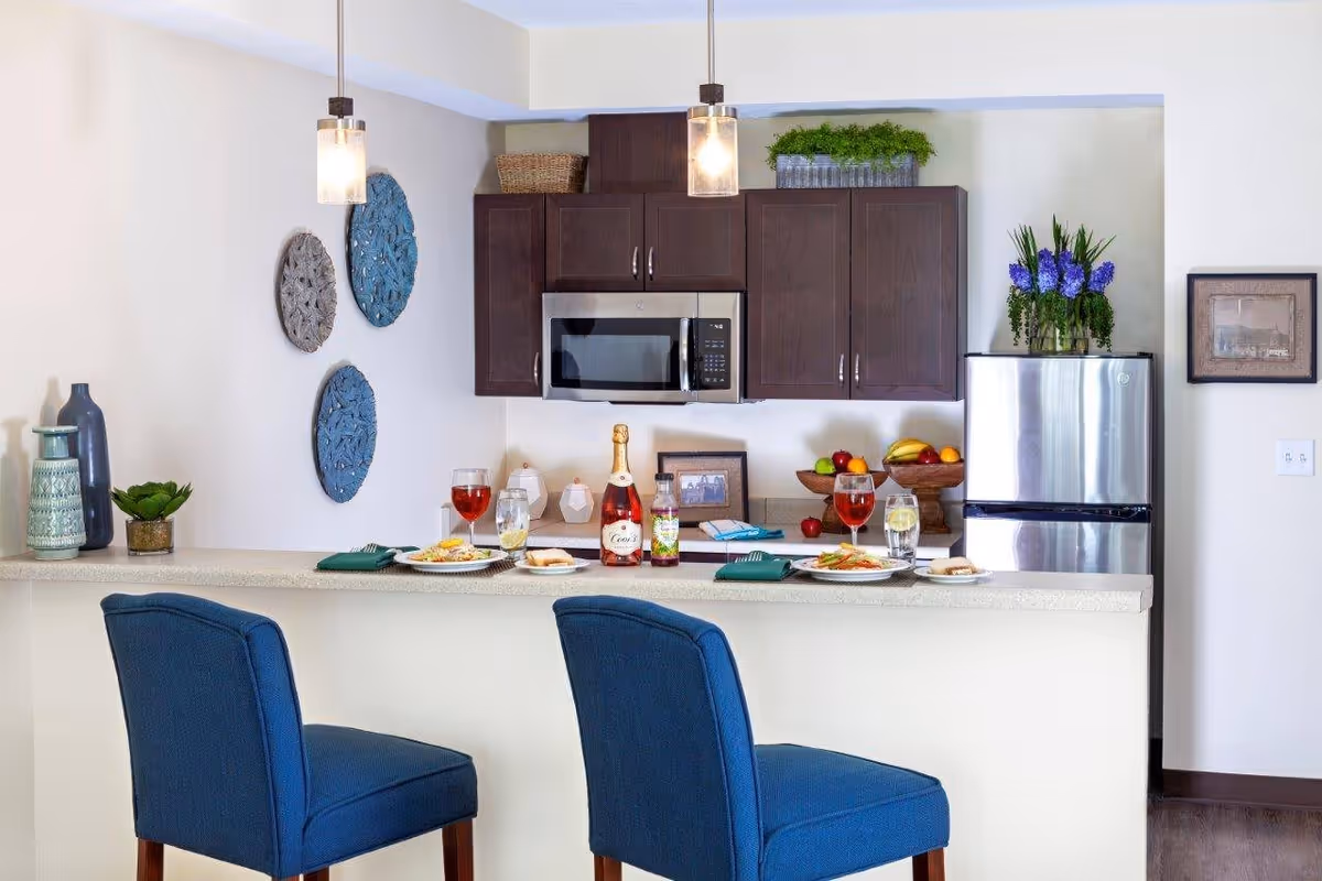 Modern kitchen area with dark wood cabinets, a stainless steel microwave and refrigerator, a countertop with two blue upholstered chairs, and a set table with plates of food, glasses of drinks, and a bottle of sparkling beverage. Decorative items include wall art, vases, and a bowl of fruit.