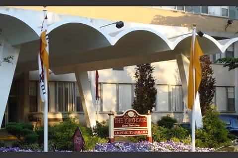Entrance of The York House Apartments featuring a distinctive white arched canopy, flags on poles, landscaped bushes and flowers, and a sign indicating luxury senior living with apartments available.