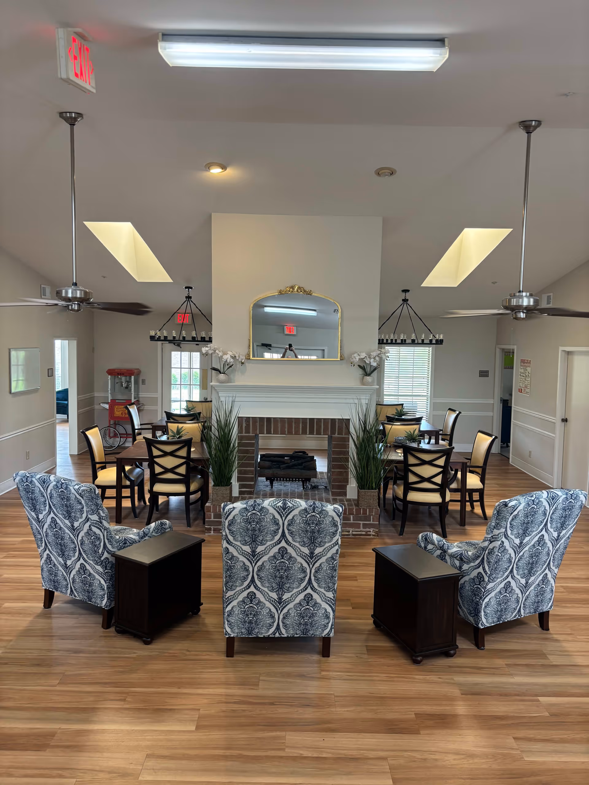 Interior view of a senior living facility common area with three patterned armchairs facing a brick fireplace with a gold-framed mirror above it. There are two ceiling fans, wooden flooring, and several dining tables with chairs in the background. Potted plants and a popcorn machine are also visible.