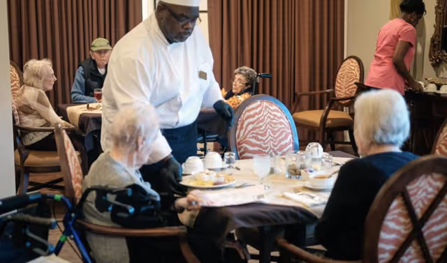A dining room in a senior living facility where elderly residents are seated at tables. A staff member dressed in a white uniform and black gloves is serving food to one of the residents. Another staff member is seen in the background attending to something on a sideboard. The room has patterned chairs and brown curtains.