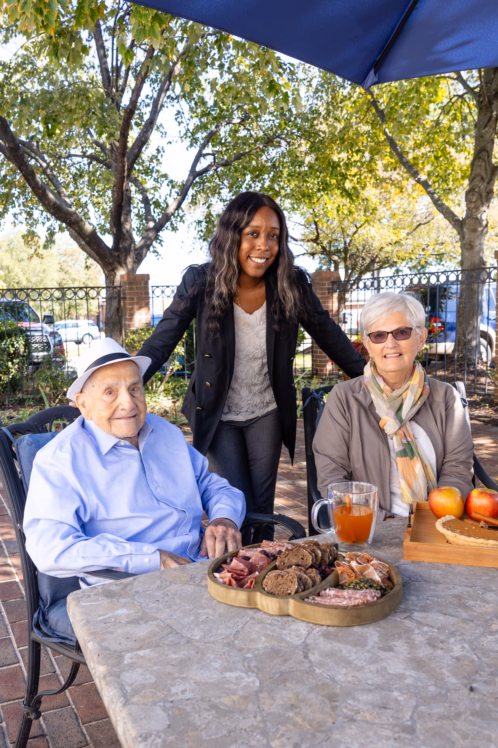 An elderly man wearing a light blue shirt and white hat and an elderly woman with white hair, glasses, and a colorful scarf sit at a stone table outdoors with a younger woman standing behind them smiling. The table has a wooden tray with apples and a pie, a glass mug with a beverage, and a divided wooden platter with various meats and crackers. Trees and a fence are visible in the background under a blue umbrella.