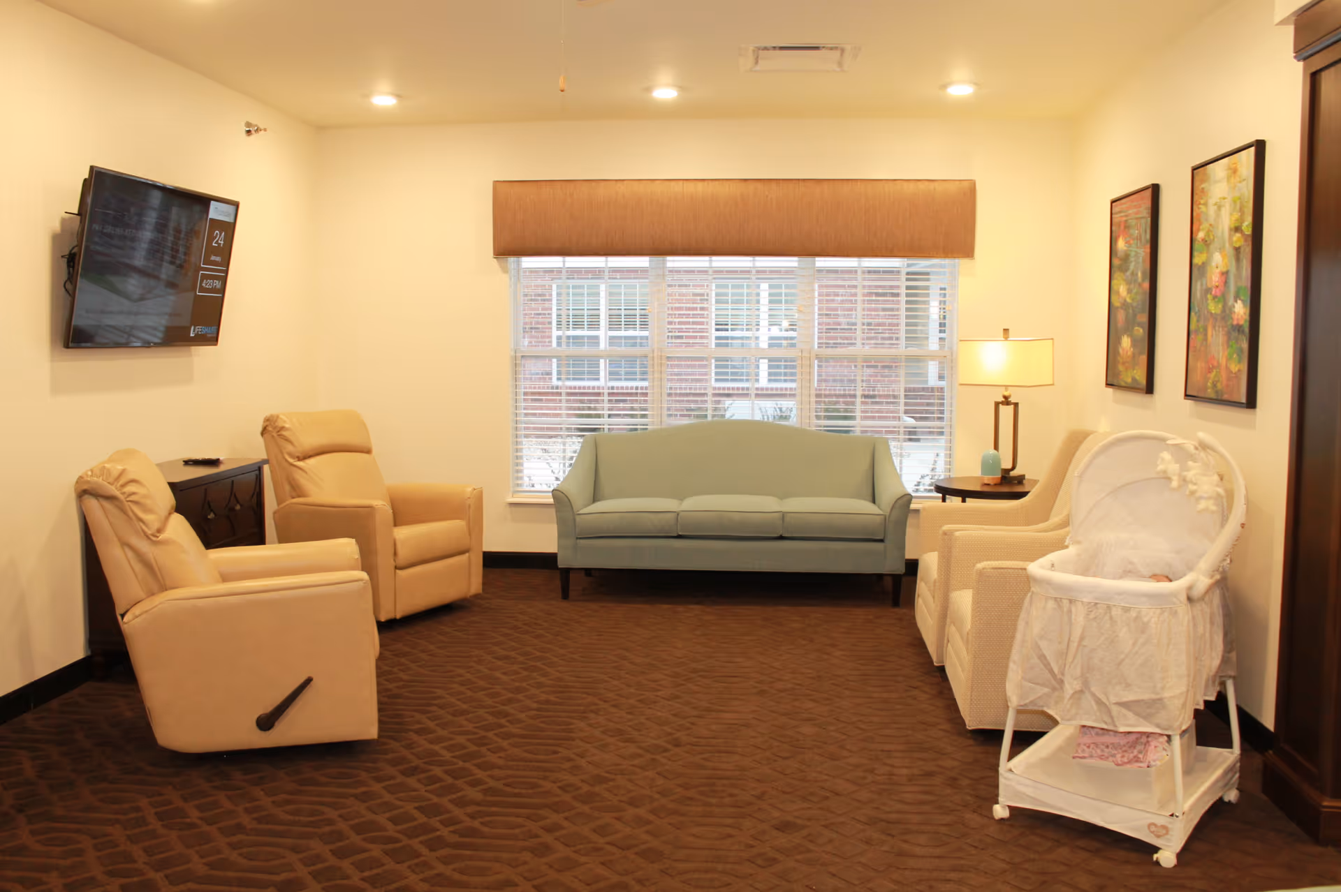 Bright common living room with a blue sofa centered under a window, two tan recliners, a wall-mounted TV, side tables with lamps, and a white bassinet.