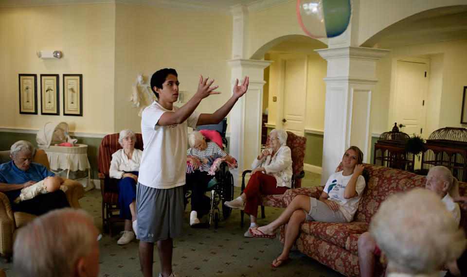A group of elderly people seated in a living room area of an assisted living facility, watching a young man in casual clothes who is standing and gesturing with his hands. The room has comfortable chairs and sofas, light-colored walls, framed artwork, and a decorative archway.