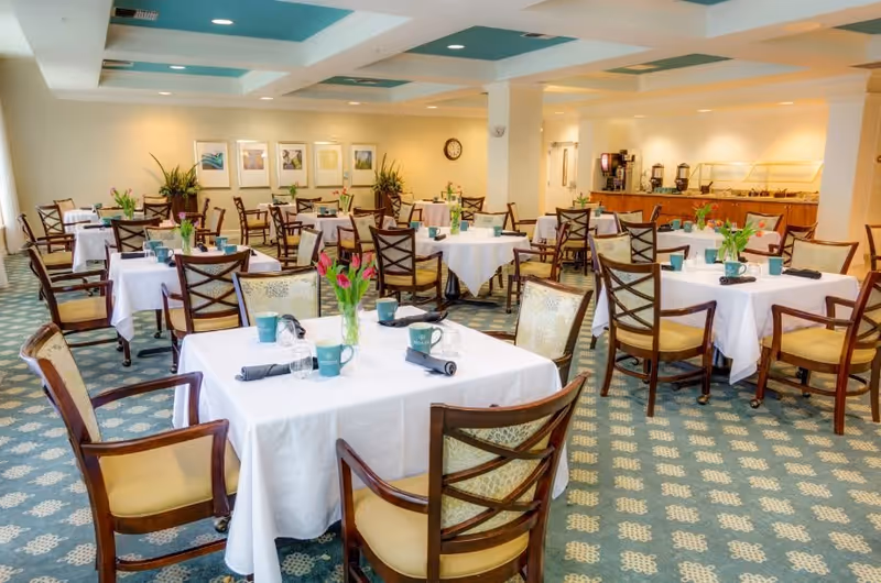 A bright and spacious dining room with multiple tables covered in white tablecloths, each set with cups, napkins, and small flower arrangements. Wooden chairs with cushioned seats surround the tables. The room has patterned carpet flooring, framed artwork on the walls, and a buffet area with coffee machines and food trays in the background.